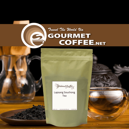 A stand-up pouch of Gourmet Coffee.net Lapsang Souchong tea stands on a wooden surface with loose tea leaves in front and blurred tea-making vessels in the background.